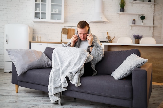 Jeune homme grippé, assis sur le canapé à la maison, enveloppé dans une couverture.