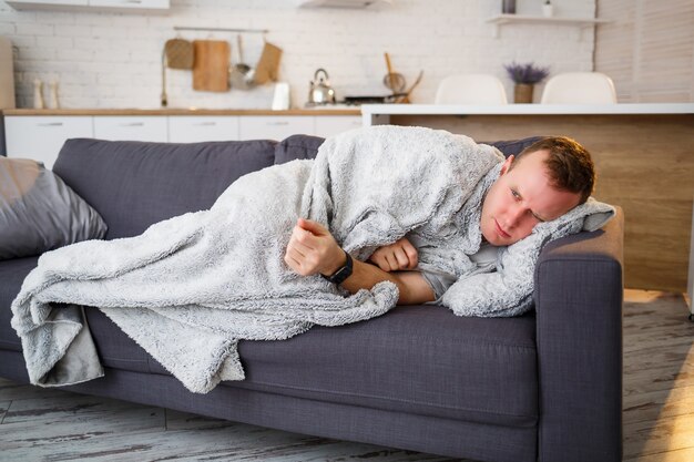 Jeune homme grippé, assis sur le canapé à la maison, enveloppé dans une couverture.