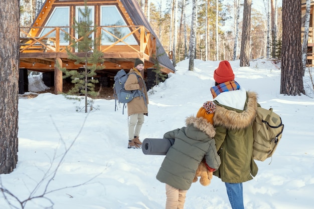 Jeune homme avec un gros sac à dos debout dans la forêt contre une maison de campagne et regardant sa femme et sa fille se diriger vers lui