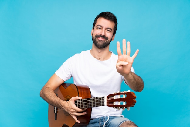 Jeune homme avec guitare sur bleu isolé heureux et comptant quatre avec les doigts