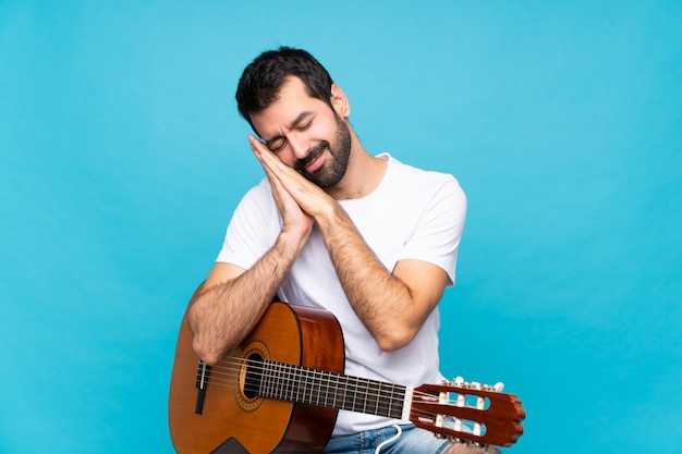 Jeune homme avec une guitare sur fond bleu isolé, geste de sommeil en expression dorable