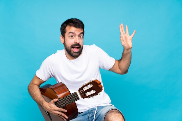 Jeune homme avec guitare sur mur bleu isolé nerveux et effrayé