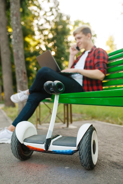 Jeune homme avec gyroboard assis sur le banc dans le parc. Loisirs de plein air avec gyroscope électrique. Transport écologique avec technologie d'équilibre, véhicule à gyroscope électrique