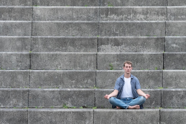 Jeune homme habillé décontracté assis dans les escaliers et méditant. Concept de santé mentale, de pleine conscience et de yoga avec espace de copie.