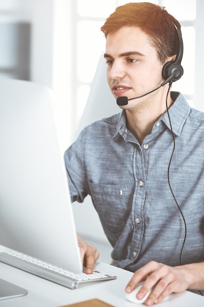 Jeune homme habillé décontracté utilisant un casque et un ordinateur tout en parlant avec des clients en ligne au bureau