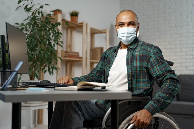 Photo jeune homme handicapé en fauteuil roulant assis à sa table de travail portant un masque facial