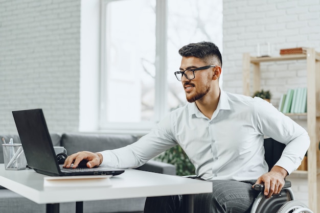 Jeune homme handicapé positif en fauteuil roulant travaillant au bureau avec ordinateur portable