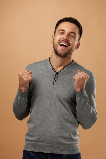 Un jeune homme heureux, beau et barbu dans un pull gris décontracté lève le poing sur un fond beige excité.