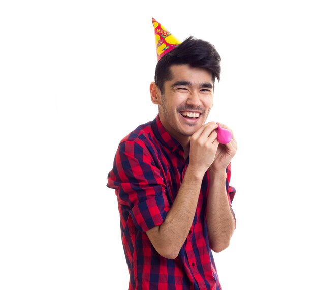 Jeune homme heureux en chemise à carreaux rouge avec un chapeau de fête soufflant un ballon rose sur fond blanc en studio