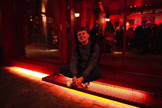 Jeune homme heureux est assis sur un bar avec une lumière scintillante au néon et des sourires.