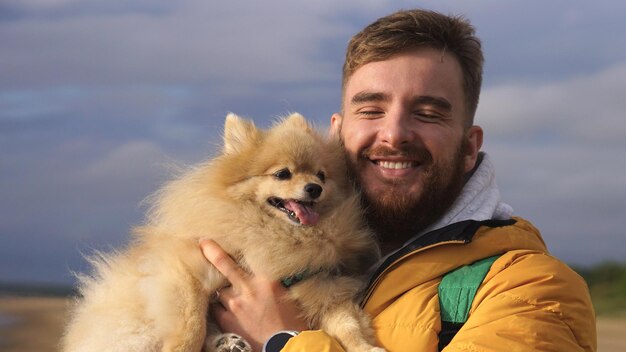 Jeune homme heureux marchant avec son chien spitz poméranien sur la plage tient le chiot sur les mains des gens