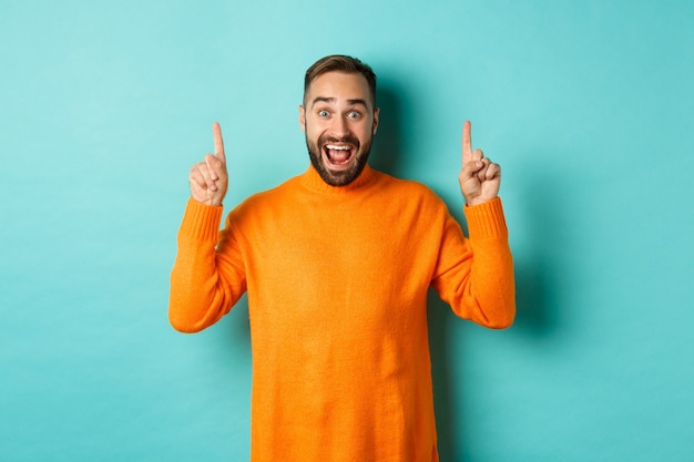 Jeune homme heureux montrant la publicité, pointant les doigts vers le haut annonçant l'offre promotionnelle, debout sur fond turquoise.