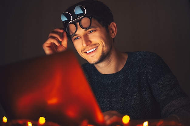 Jeune homme heureux portant des lunettes rondes, à la recherche d'un ordinateur portable dans une pièce sombre avec des guirlandes à la maison.