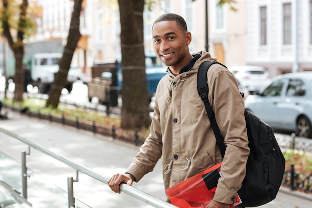 Jeune homme heureux portant un sac à dos debout dans la rue tout en tenant un livre et en regardant à l'avant