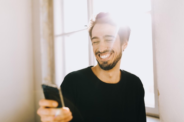 Un jeune homme heureux qui utilise son téléphone près de la fenêtre de sa maison.