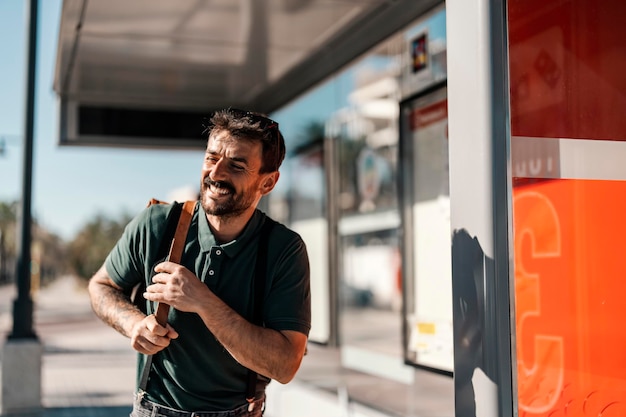 Un jeune homme heureux se tient à l'arrêt de bus et attend un bus