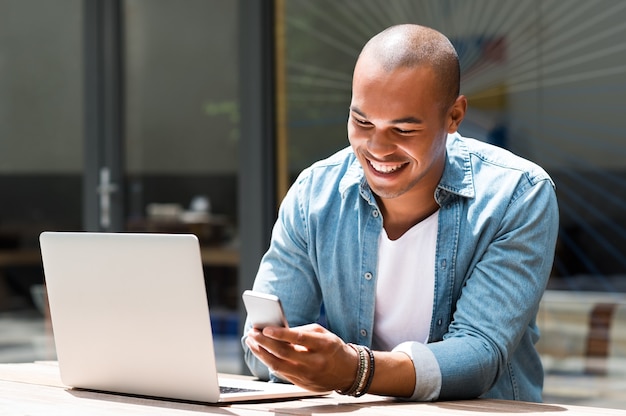 Jeune homme heureux souriant et tenant un smartphone assis en plein air avec ordinateur portable