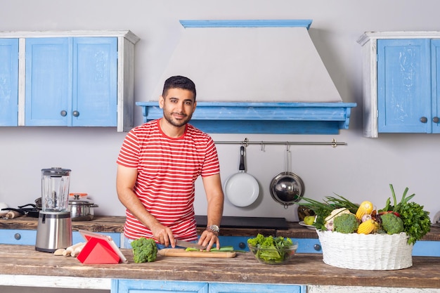 Jeune homme heureux en t-shirt décontracté coupant des légumes, préparant une salade végétarienne dans une cuisine moderne, cuisinant des légumes verts sur la table, de la nourriture végétalienne, une nutrition diététique et regardant la caméra en souriant