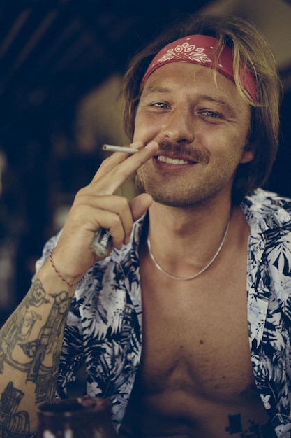 Un jeune homme hippie beau dans un bandana rouge avec un torse nu roule une cigarette de tabac dans un café à Bali.