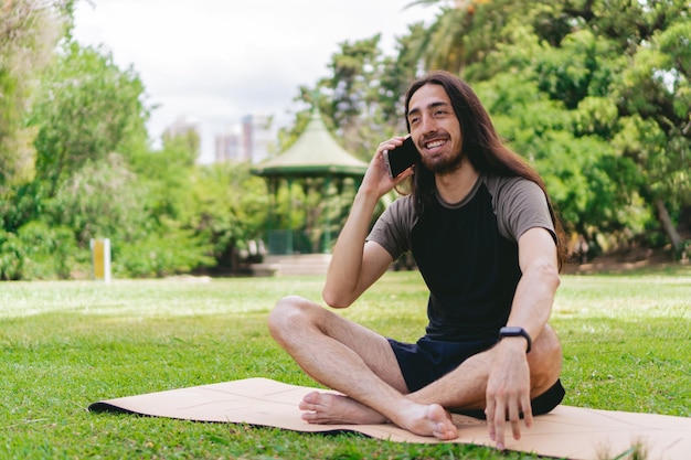 Jeune homme hippie latin assis dans la position du lotus sur un tapis de yoga parlant au téléphone dans un champ ouvert avec un belvédère derrière