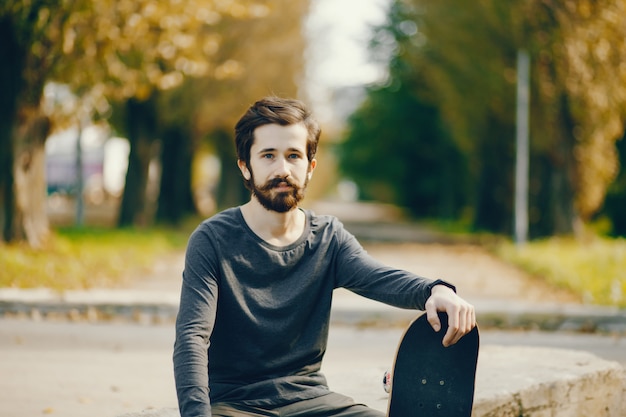 jeune homme hipster assis dans un parc ensoleillé d&#39;été avec une planche à roulettes dans ses mains