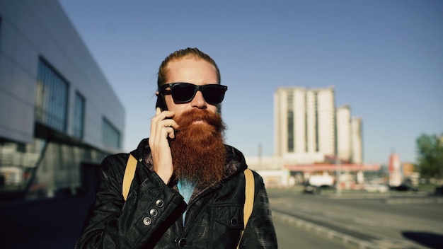 Jeune homme hipster barbu à lunettes de soleil souriant et parlant de smartphone tout en voyageant dans la rue de la ville