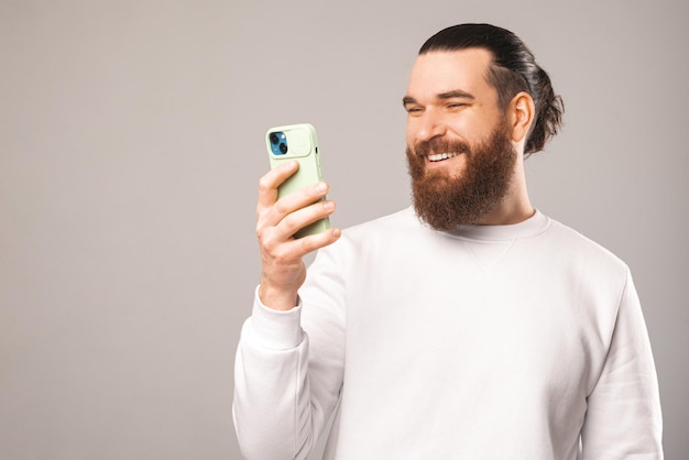 Jeune homme hipster barbu souriant tient et utilise son smartphone