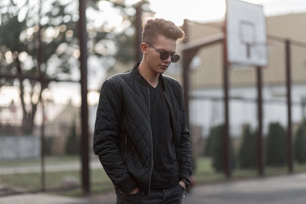 Jeune homme hipster beau mode avec des lunettes de soleil élégantes en veste tendance noire se dresse dans un stade de la rue