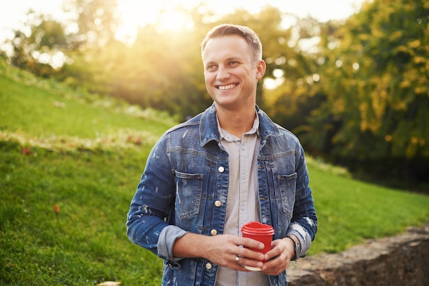 Jeune homme Hipster debout avec du café à emporter dans le parc, souriant agréablement à la caméra. Heureux beau mec sans soucis en veste en jean bleu