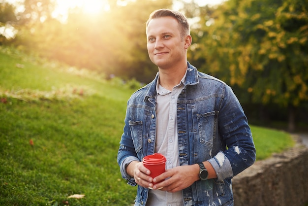 Jeune homme Hipster debout avec du café à emporter dans le parc, souriant agréablement à la caméra. Heureux beau mec sans soucis en veste en jean bleu