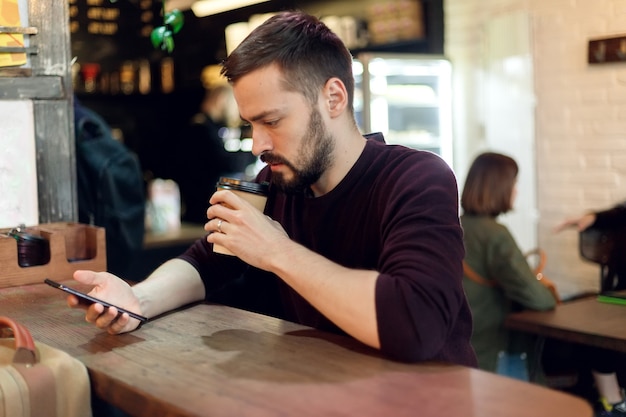 Jeune homme hipster envoyant des SMS avec son téléphone portable au bar et prenant un cappuccino. Jeune homme à la mode / hipster buvant du café expresso au café de la ville pendant l'heure du déjeuner et travaillant sur une tablette