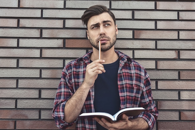 Jeune homme hipster portant des lunettes lisant un livre ou un carnet de notes prendre des notes et des idées sur fond de mur de brique.