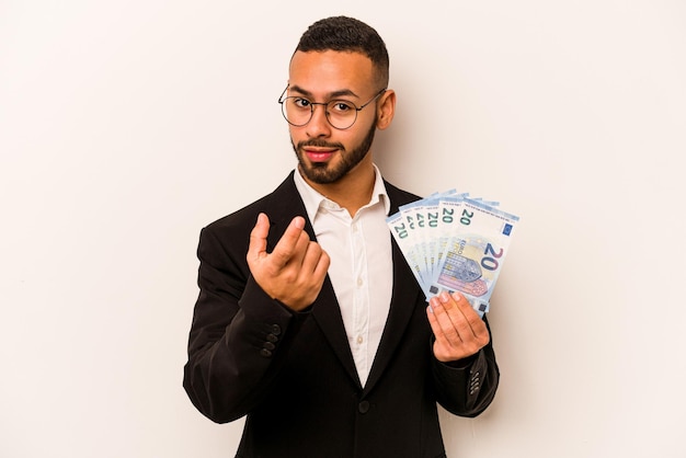 Jeune homme hispanique d'affaires tenant des billets de banque isolés sur fond blanc pointant du doigt vers vous comme s'il vous invitait à vous rapprocher