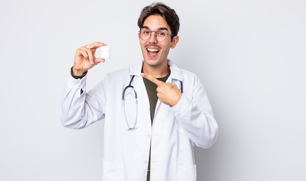 Jeune homme hispanique à l'air excité et surpris en pointant sur le côté. médecin avec concept de bouteille de pilules