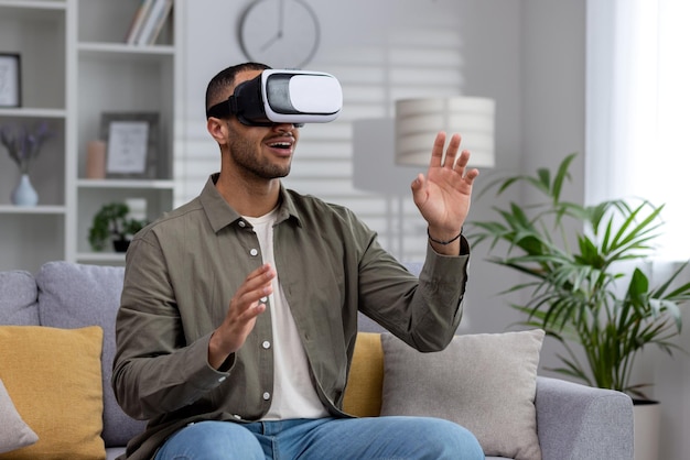 Jeune homme hispanique assis sur un canapé à la maison portant des lunettes de réalité virtuelle joue une vidéo en ligne