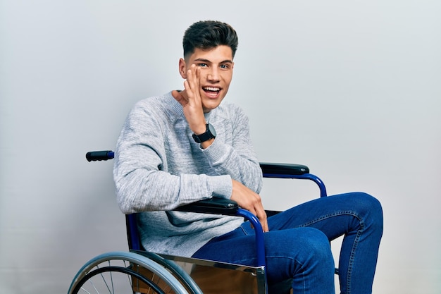 Jeune homme hispanique assis sur un fauteuil roulant main sur la bouche racontant une rumeur secrète, chuchotant une conversation malveillante