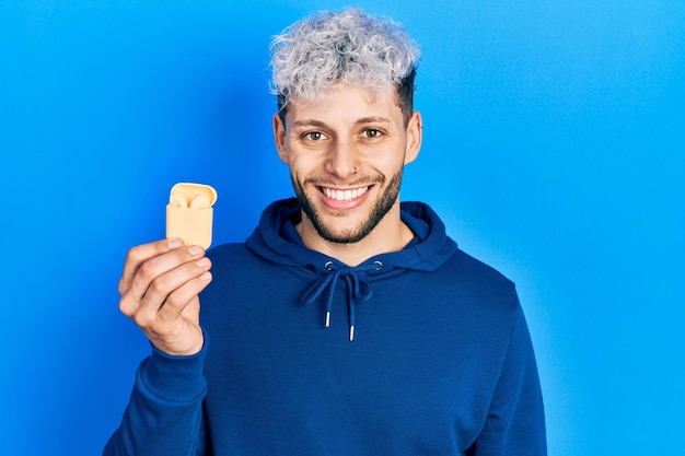 Jeune homme hispanique aux cheveux teints modernes tenant des écouteurs sans fil à l'air positif et heureux debout et souriant avec un sourire confiant montrant les dents