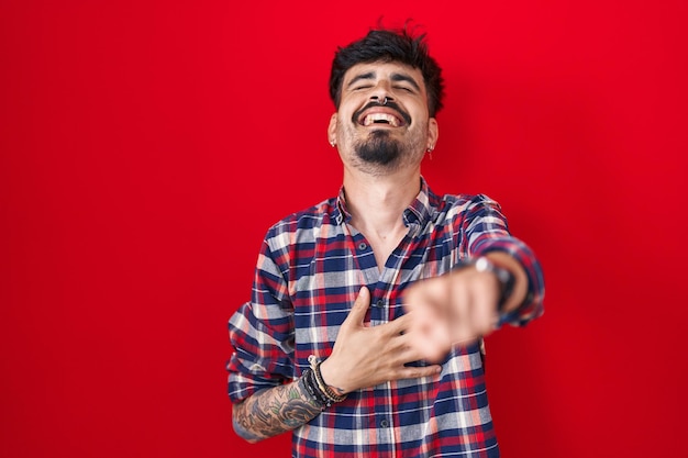 Jeune homme hispanique avec barbe debout sur fond rouge riant de vous pointant le doigt vers la caméra avec la main sur l'expression de la honte du corps