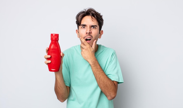 Jeune homme hispanique avec la bouche et les yeux grands ouverts et la main sur le concept de ketchup au menton