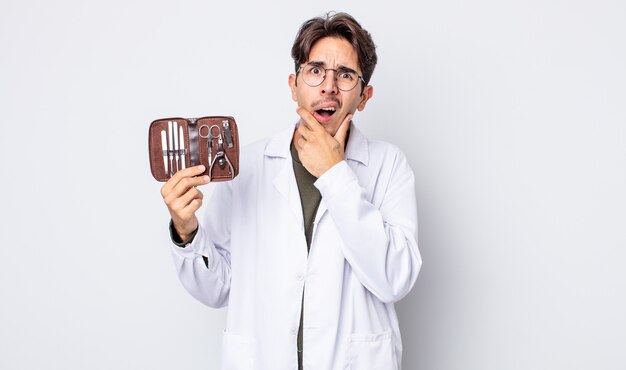 Jeune homme hispanique avec la bouche et les yeux grands ouverts et la main sur le menton. outils d'ongles de podologue