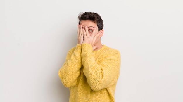 Jeune homme hispanique couvrant le visage avec les mains furtivement entre les doigts avec une expression surprise et regardant sur le côté