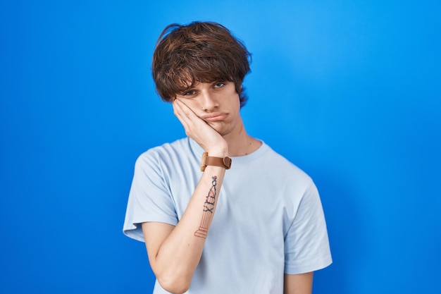 Jeune homme hispanique debout sur fond bleu pensant avoir l'air fatigué et ennuyé par des problèmes de dépression avec les bras croisés.