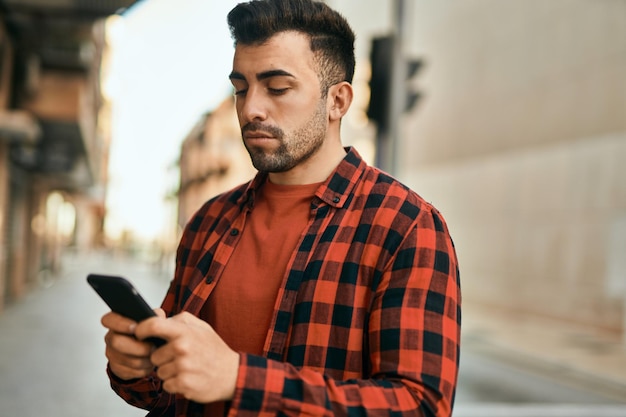 Jeune homme hispanique avec une expression sérieuse à l'aide de smartphone à la ville.