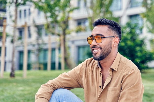 Jeune homme hispanique heureux pensant assis au parc homme latin portant des lunettes de soleil