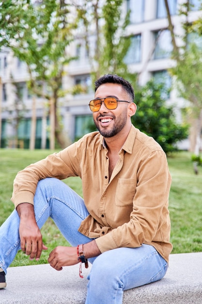 Photo jeune homme hispanique heureux pensant assis au parc homme latin portant des lunettes de soleil