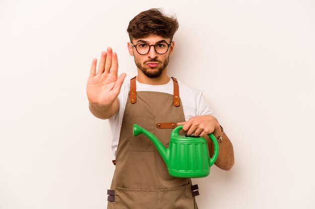 Jeune homme hispanique jardinier tenant un arrosoir isolé sur fond blanc debout avec la main tendue montrant le panneau d'arrêt vous empêchant