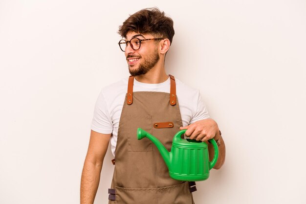 Jeune homme hispanique jardinier tenant un arrosoir isolé sur fond blanc regarde de côté souriant gai et agréable