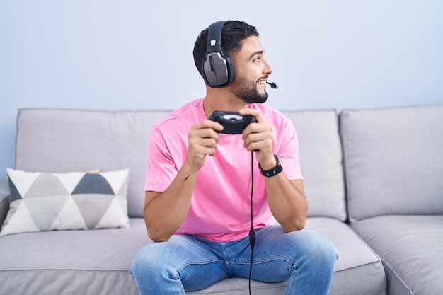 Un jeune homme hispanique jouant à un jeu vidéo tenant un contrôleur assis sur le canapé à la recherche d'un côté relaxant pose de profil avec un visage naturel et un sourire confiant