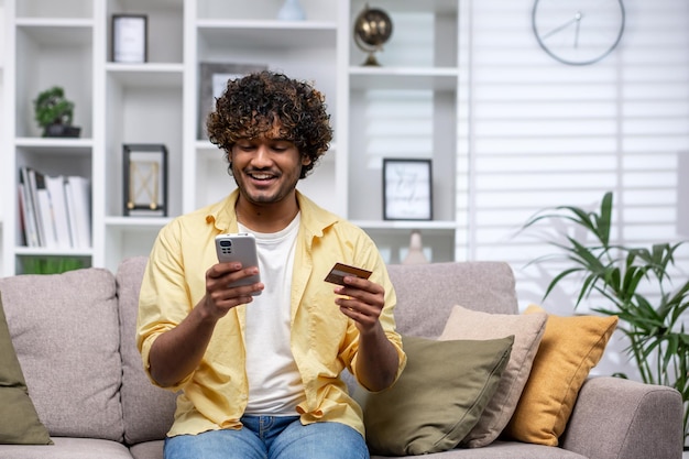 Jeune homme hispanique à la maison seul sur un canapé dans le salon assis souriant et heureux homme utilisant le téléphone