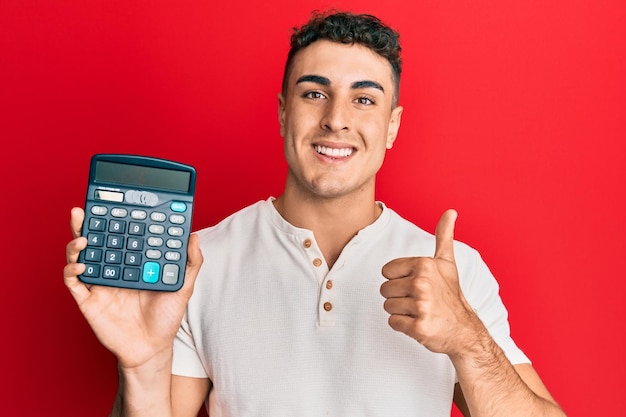 Jeune homme hispanique montrant un appareil calculatrice souriant heureux et positif pouce vers le haut faisant un excellent signe d'approbation